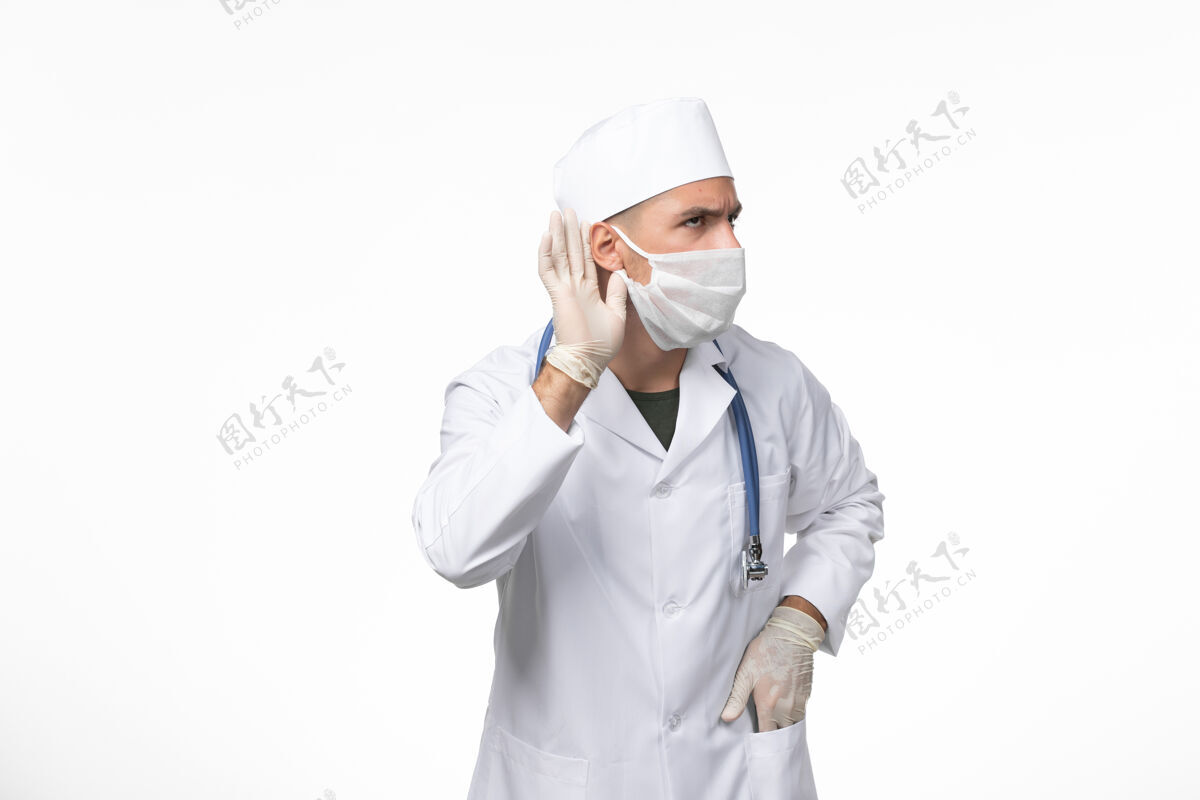 听力正面图男医生身着医疗服 戴着防毒面具 用蓝色听诊器试图听到白墙上的冠状病毒病大流行病毒正面视图面罩