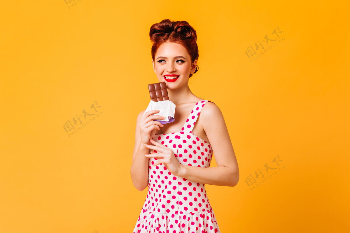 老式微笑着吃巧克力的姜汁女孩摄影棚拍摄了黄色空间中穿着圆点连衣裙的女人黑发漂亮发型