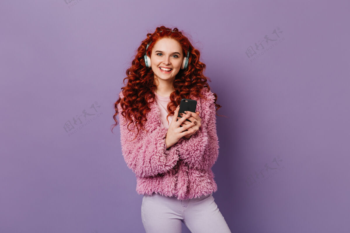 太阳镜蓝眼睛甜美的女孩微笑着看着紫色空间里的相机卷曲的红发女人穿着粉色夹克 手里拿着电话 戴着耳机摆姿势蓝色眼睛性感魅力