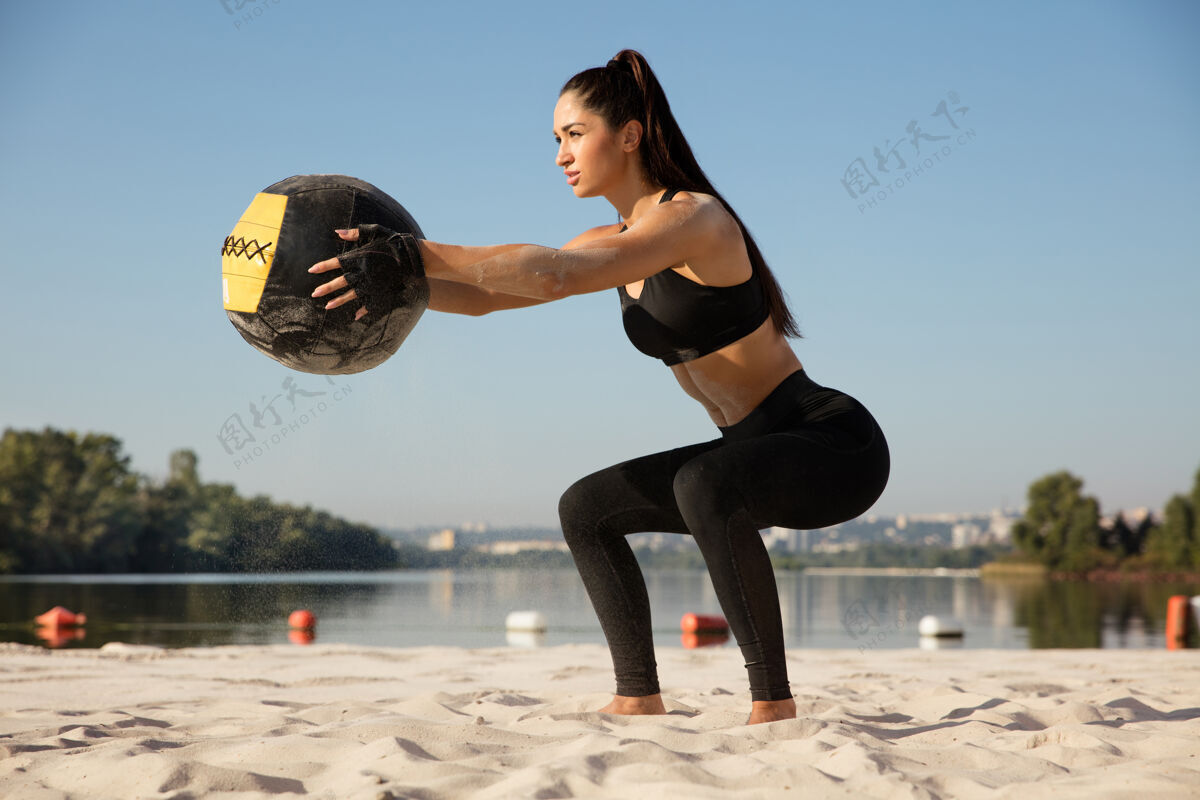 教练年轻健康的女人在沙滩上拿着球蹲着运动员空气锻炼