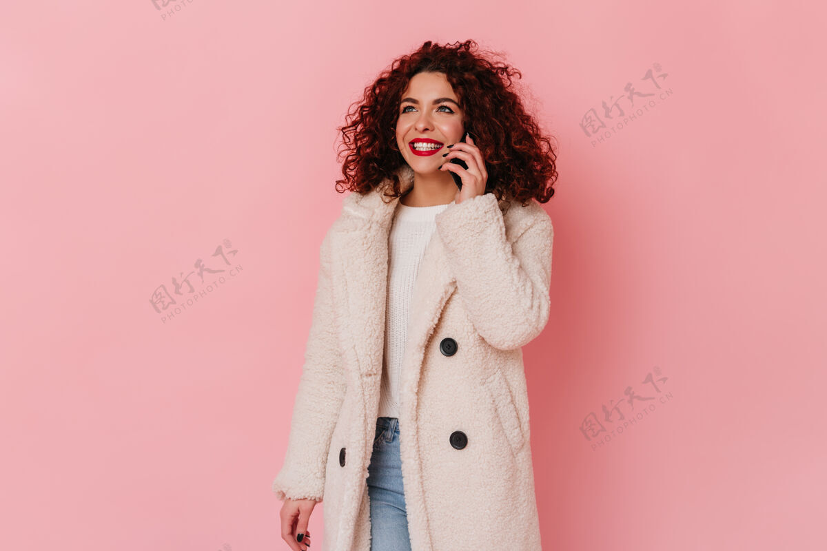 黑发穿着环保外套和牛仔裤的时髦女孩在打电话红唇白笑的女人在粉色空间摆姿势发型时尚女性