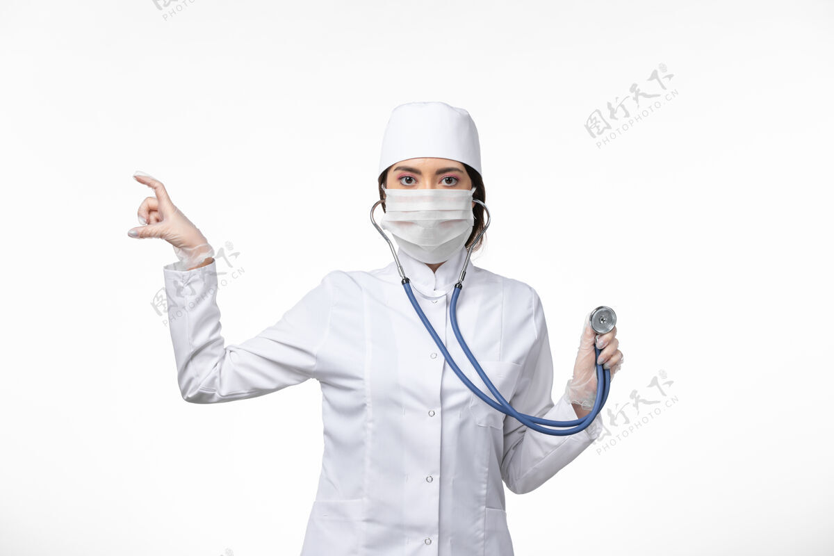 冠状病毒正面图女医生身穿白色无菌医疗服 戴口罩因冠状病毒-使用听诊器对白墙病病毒冠状病毒-大流行性疾病佩戴冠状病毒预防病毒