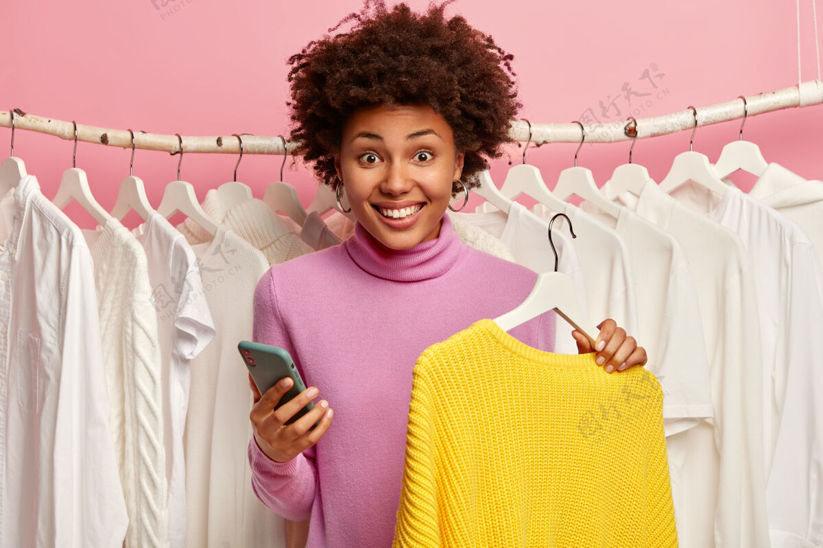 毛衣微笑的非洲头发女人 使用手机应用程序在线支付 购买新的黄色毛衣 站在衣架后面高兴女士商店