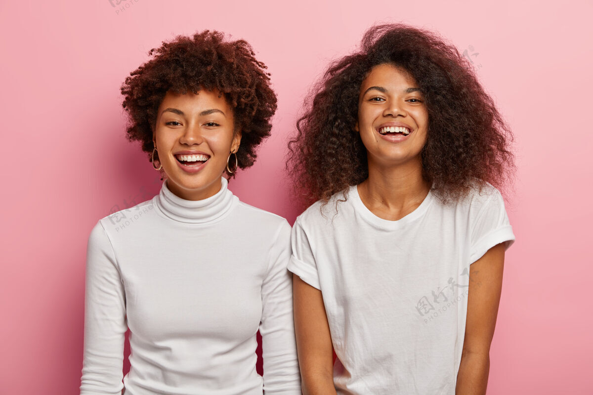 非洲摄影棚拍摄的快乐的美国黑人女姐妹们享受美好的时刻 穿着白色休闲服 笑容灿烂 在空闲时间一起玩得很开心 隔着粉色的墙关系非洲休闲