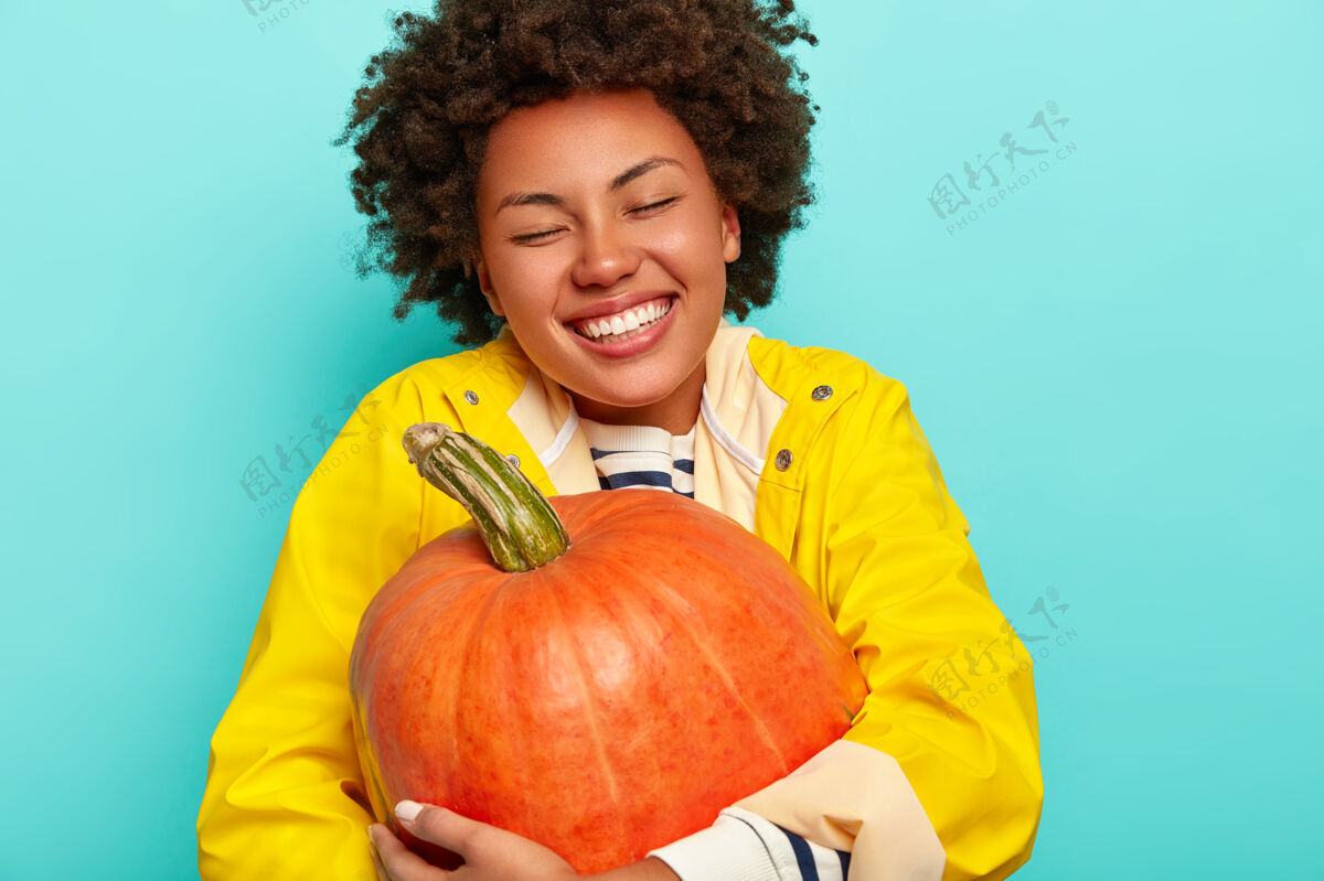 一个快乐的黑皮肤女人拥抱南瓜的照片 有牙齿的微笑 穿着黄色雨衣 感到高兴和放松 在蓝色的背景上摆姿势秋天欢呼女孩