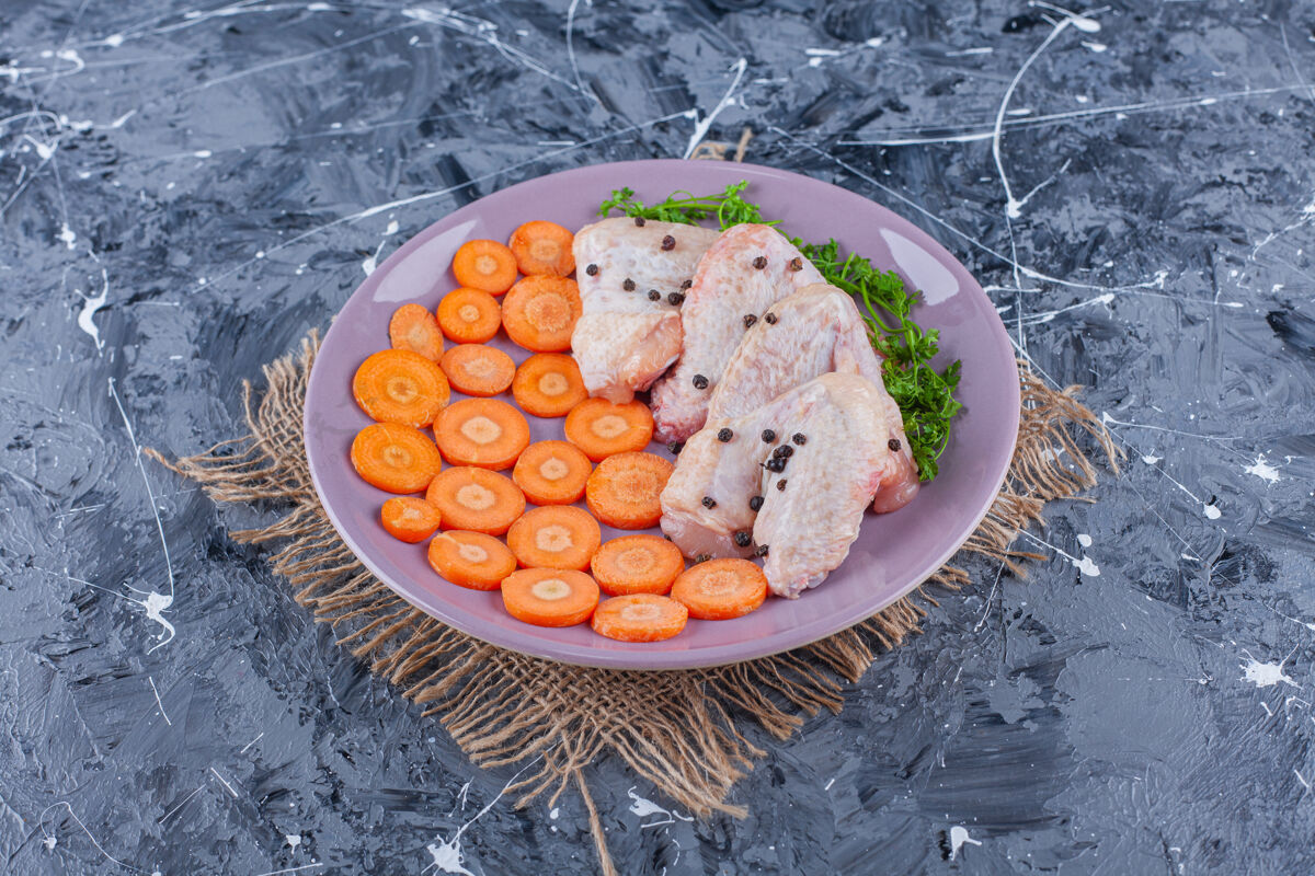 配料把切成片的胡萝卜 鸡翅和青菜放在盘子里 放在蓝色表面的粗麻布上新鲜有机鸡肉