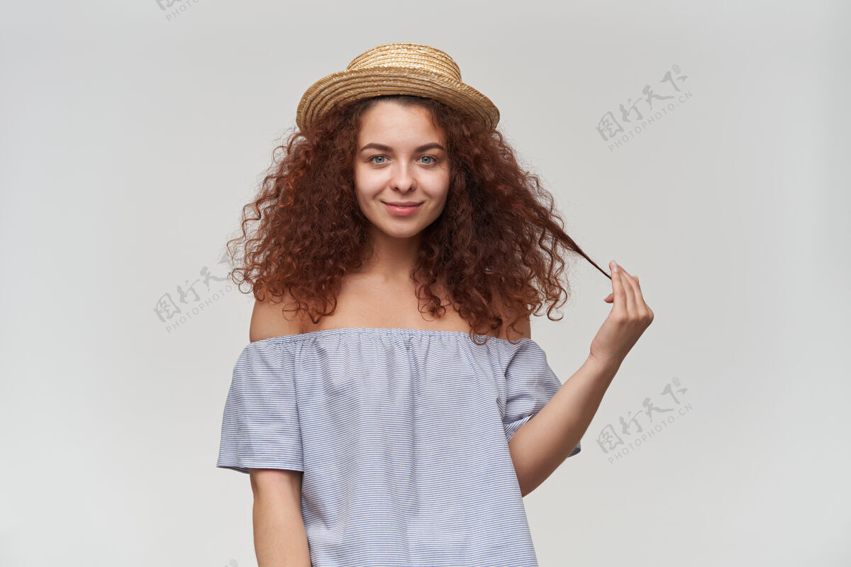 女性十几岁的女孩 快乐的女人 卷曲的姜黄色头发穿着条纹露肩衬衫和帽子玩着一缕头发 微笑隔着白色的墙年轻室内肩膀