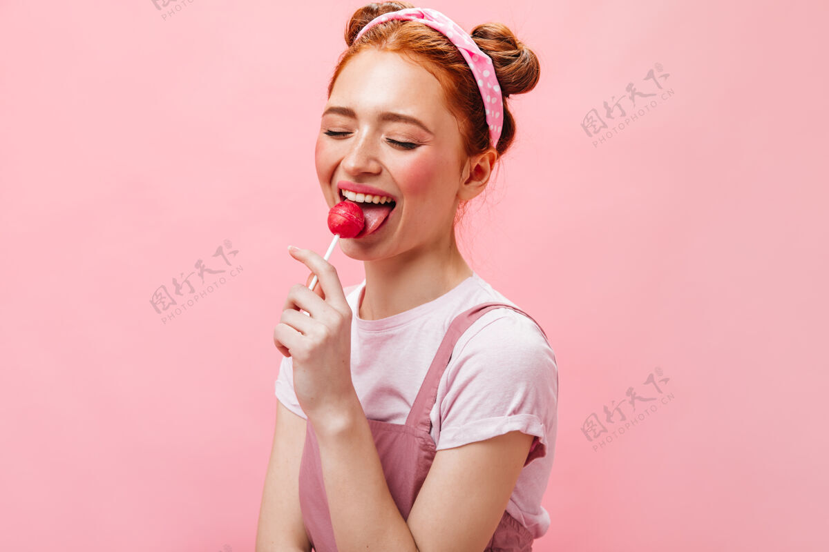 眼睛可爱的红发女人穿着白色上衣和裙子舔着粉色背景上的棒棒糖化妆品波浪发糖果