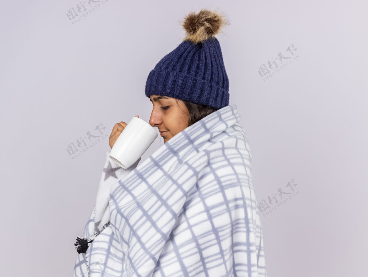 围巾站在侧视图年轻的生病的女孩戴着冬天的帽子和围巾包裹在格子呢举行杯子冬天帽子