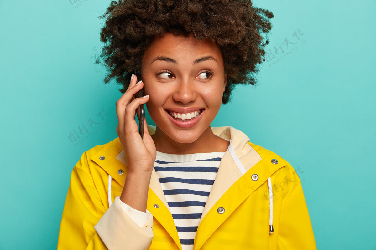 交谈卷发女人的特写镜头有着幸福的表情 打电话给好友讲着搞笑的故事 穿着黄色防水雨衣 聚精会神地站在一旁 站在蓝色背景下的室内欢呼微笑肖像
