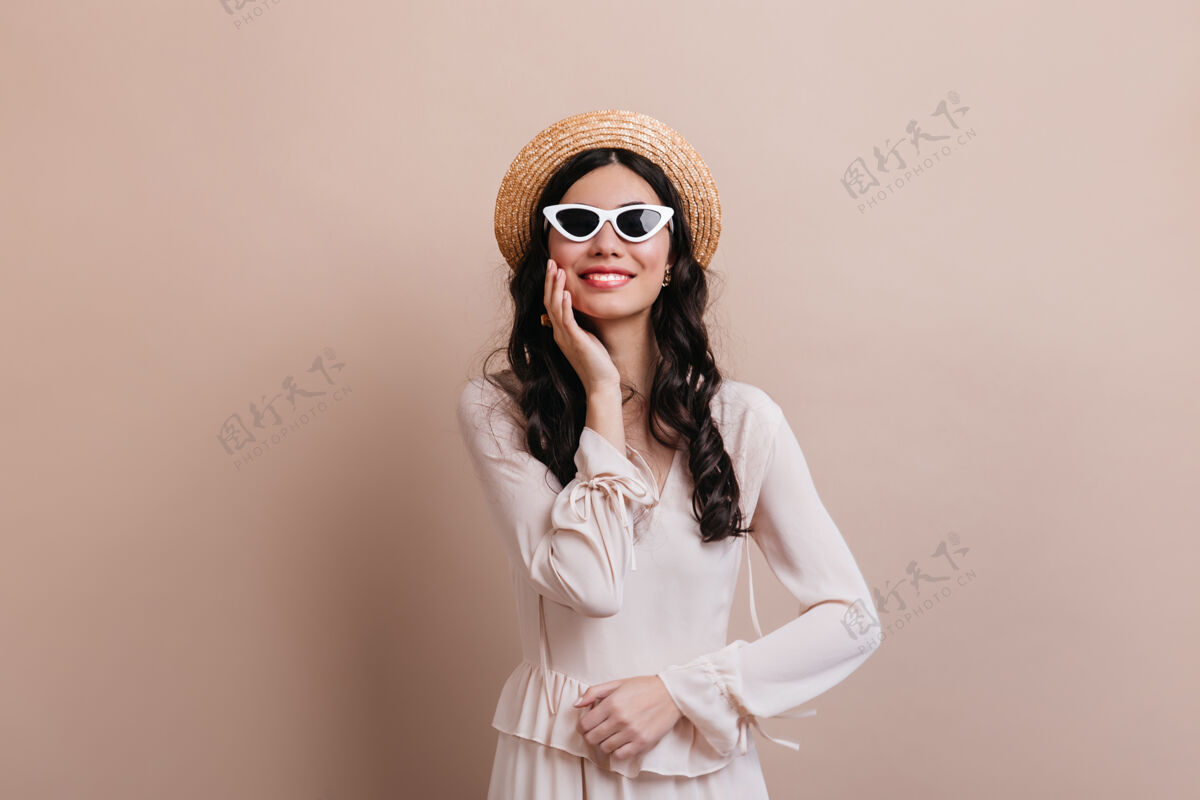 工作室拍摄戴着墨镜的迷人女人在米色背景下欢笑戴着草帽的快乐黑发女人的正面视图时尚无忧无虑配饰