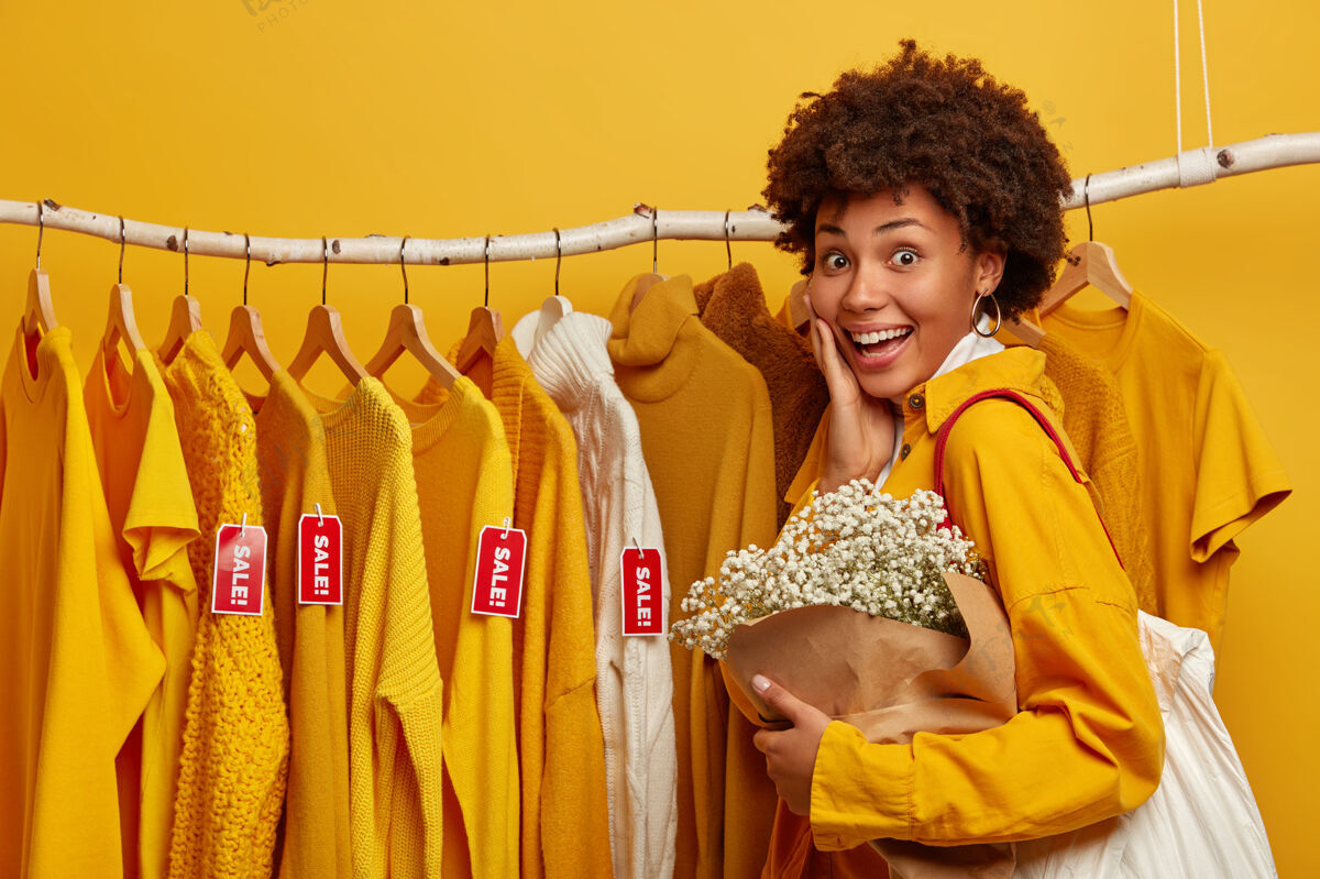 欣喜若狂快乐的卷发女买家选择挂在衣架上出售的衣服 拎着包 摆着花束 隔离在黄色背景上非洲消费主义零售