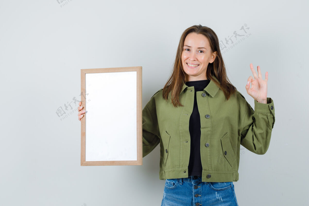 休闲身穿绿色夹克的年轻女子手持空白镜框 摆出“ok”的姿势 正面朝上看秀积极手势
