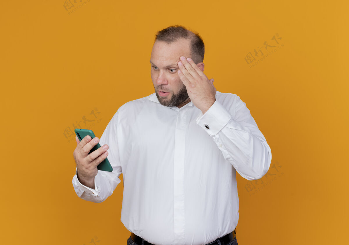 胡子一个留着胡子 身穿白衬衫的男人站在橘色的墙上 看着智能手机的屏幕 感到既不高兴又困惑穿男人看