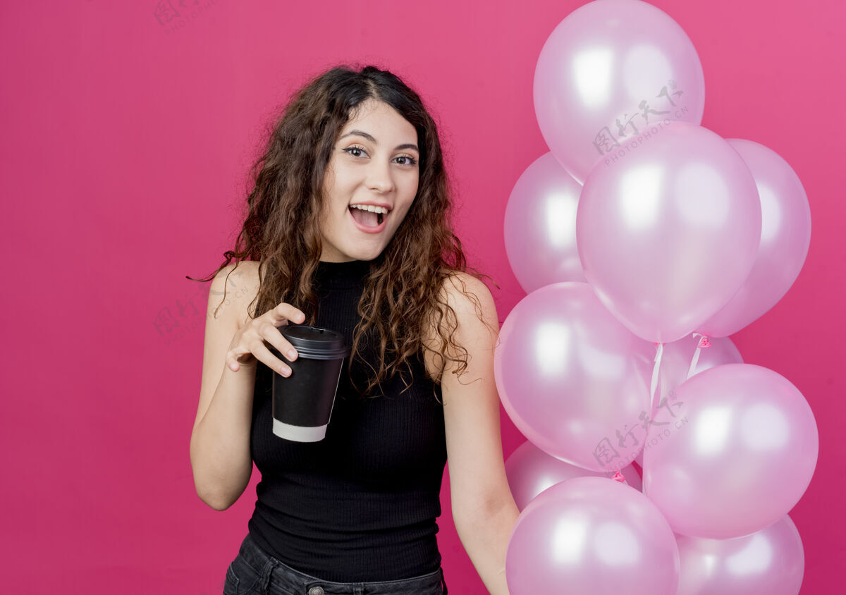 空气一个年轻漂亮的女人 卷发 手里拿着一束气球和咖啡杯 站在粉红色的墙上 开心地笑着年轻抱着卷发