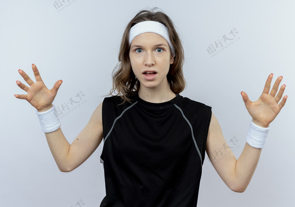 运动装身穿黑色运动服 头戴头巾 双臂高举站在白墙上的年轻健身女孩看站手臂