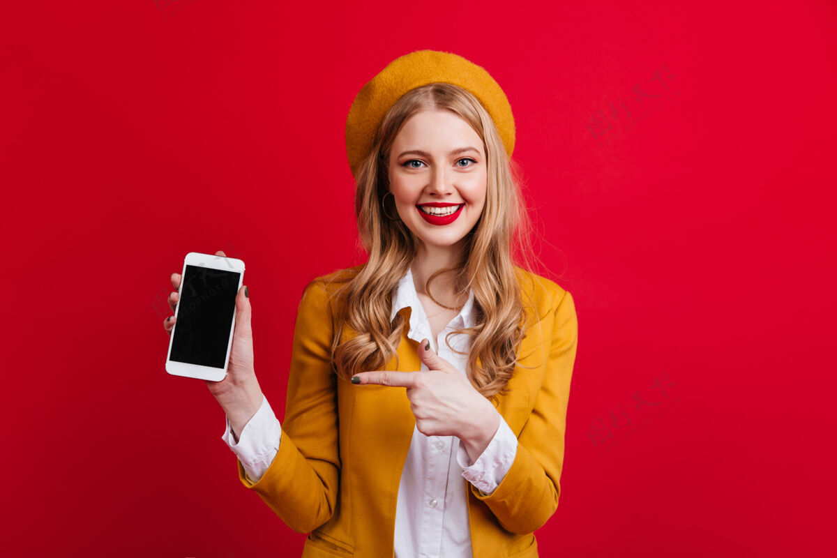 表情迷人的法国女子手持智能手机 屏幕空白戴黄色贝雷帽的女孩正对着数码设备指指点点帽子空白屏幕微笑