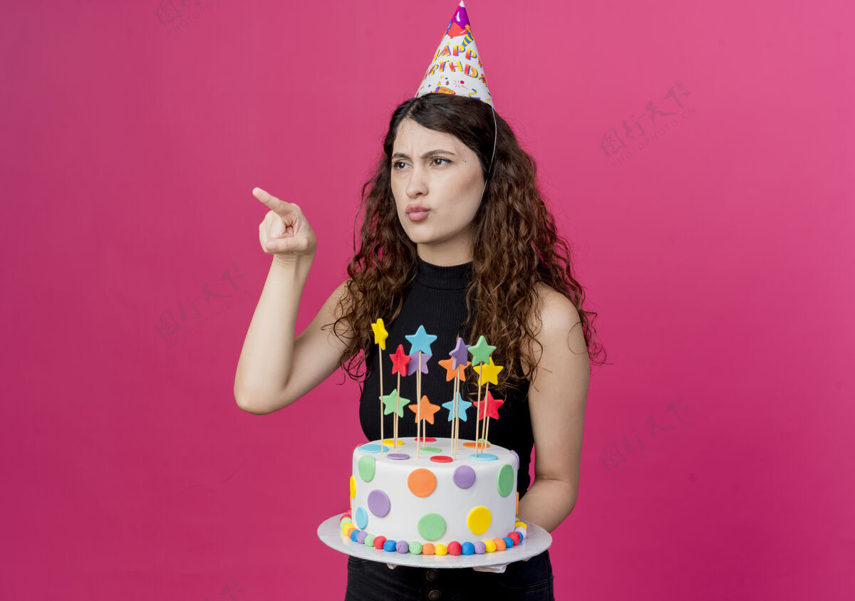 手指戴着节日帽的卷发美女拿着生日蛋糕 手指指着站在粉色墙上的某个看起来不高兴的生日派对概念卷发拿着不高兴