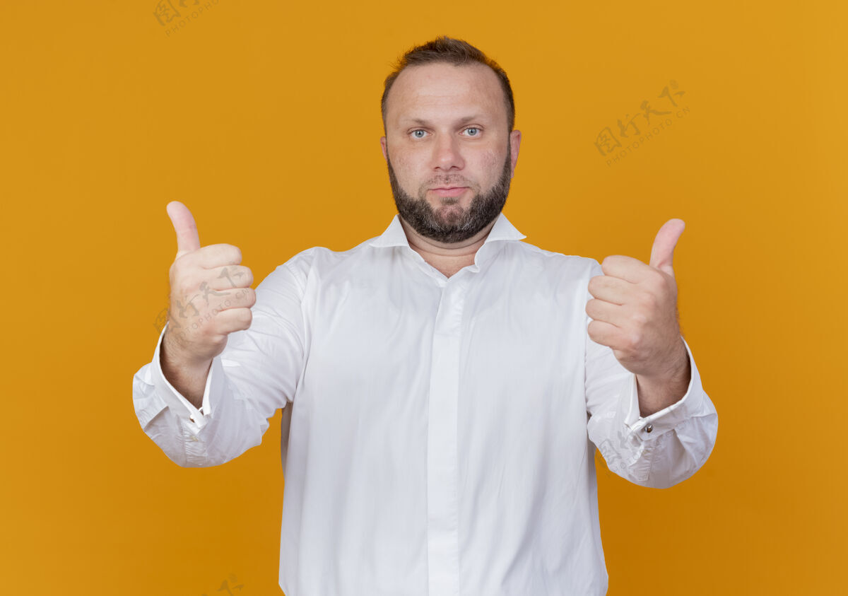 穿着满脸胡须的男人穿着白衬衫 自信地站在橙色的墙上 竖起大拇指站立展示自信