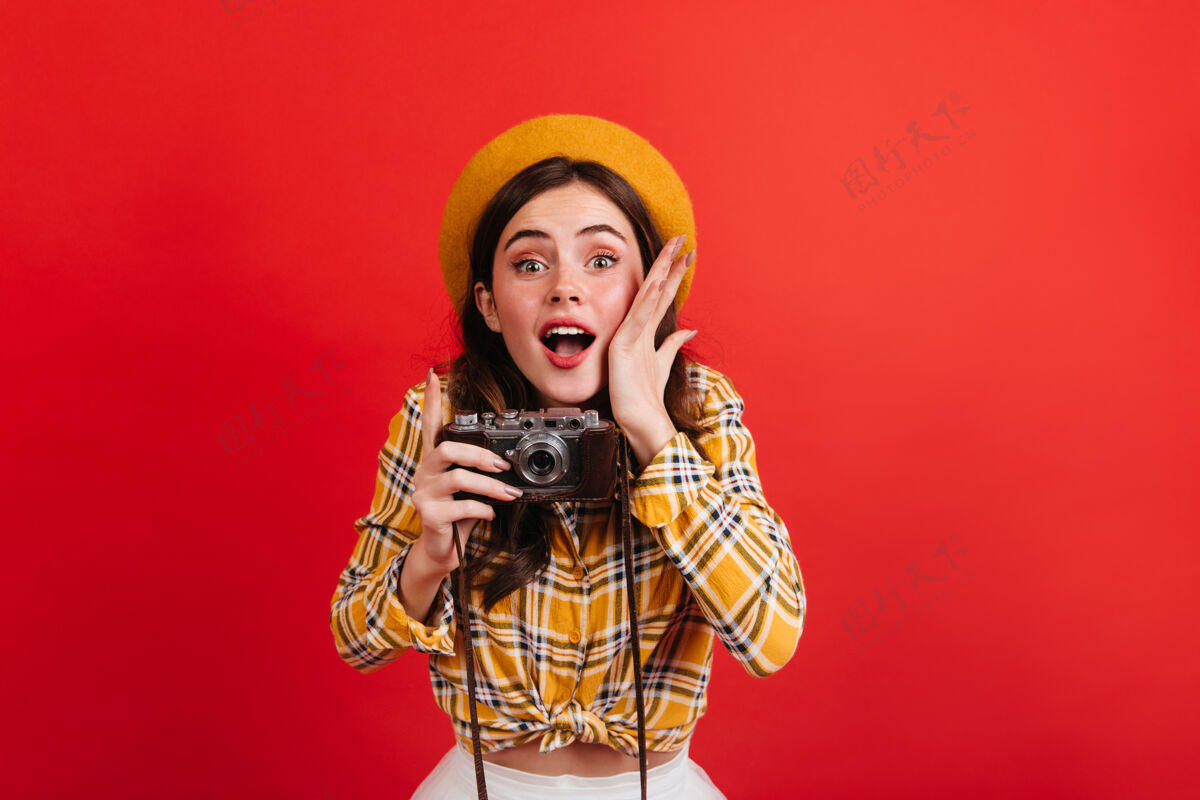 法国美丽的年轻女孩博主在复古相机上拍照红墙上有一个穿着橘色衣服戴着帽子的绿眼睛女人的肖像室内明亮帽子