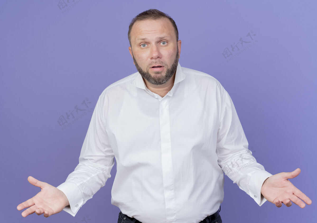 看一个留着胡须 穿着白衬衫的男人站在蓝色的墙上 看起来困惑不解地伸出双臂 好像在问或有疑问站男人困惑