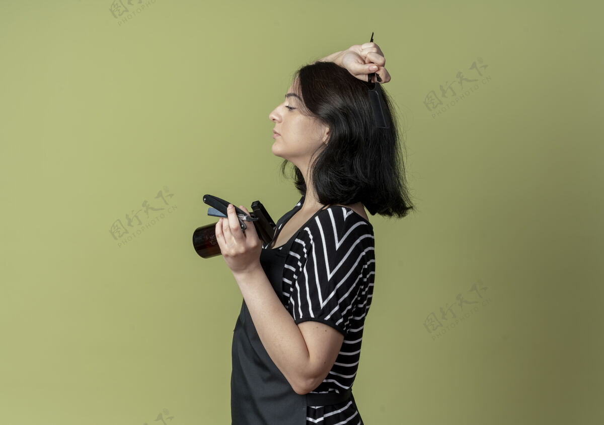 年轻年轻漂亮的女理发师围着围裙 手里拿着喷雾梳和剃刀 站在明亮的墙上光美发师剃须刀