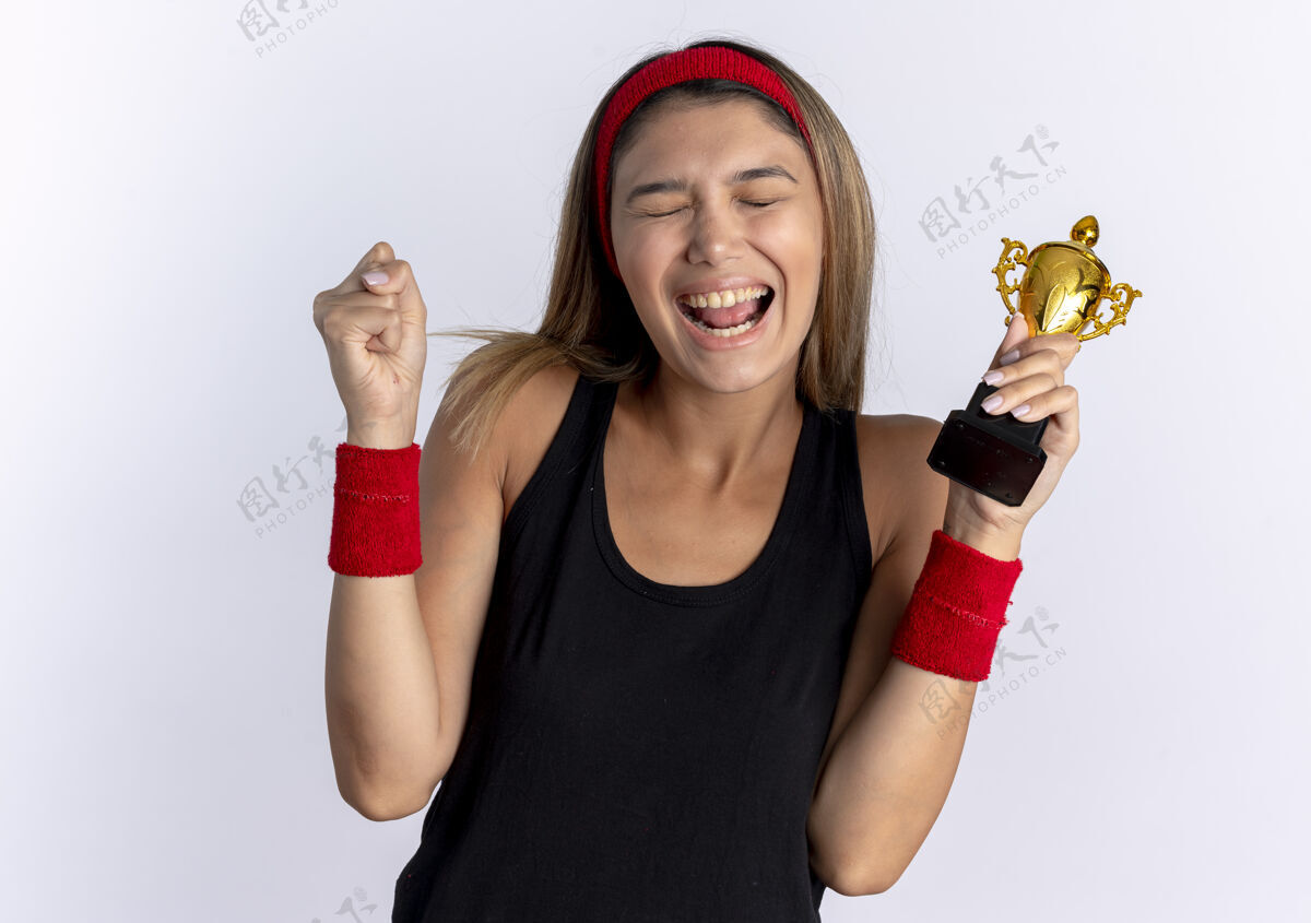 运动装身穿黑色运动服 头戴红色头巾 手持奖杯 握紧拳头的年轻健身女孩站在白墙上 快乐而兴奋站着女孩抱着
