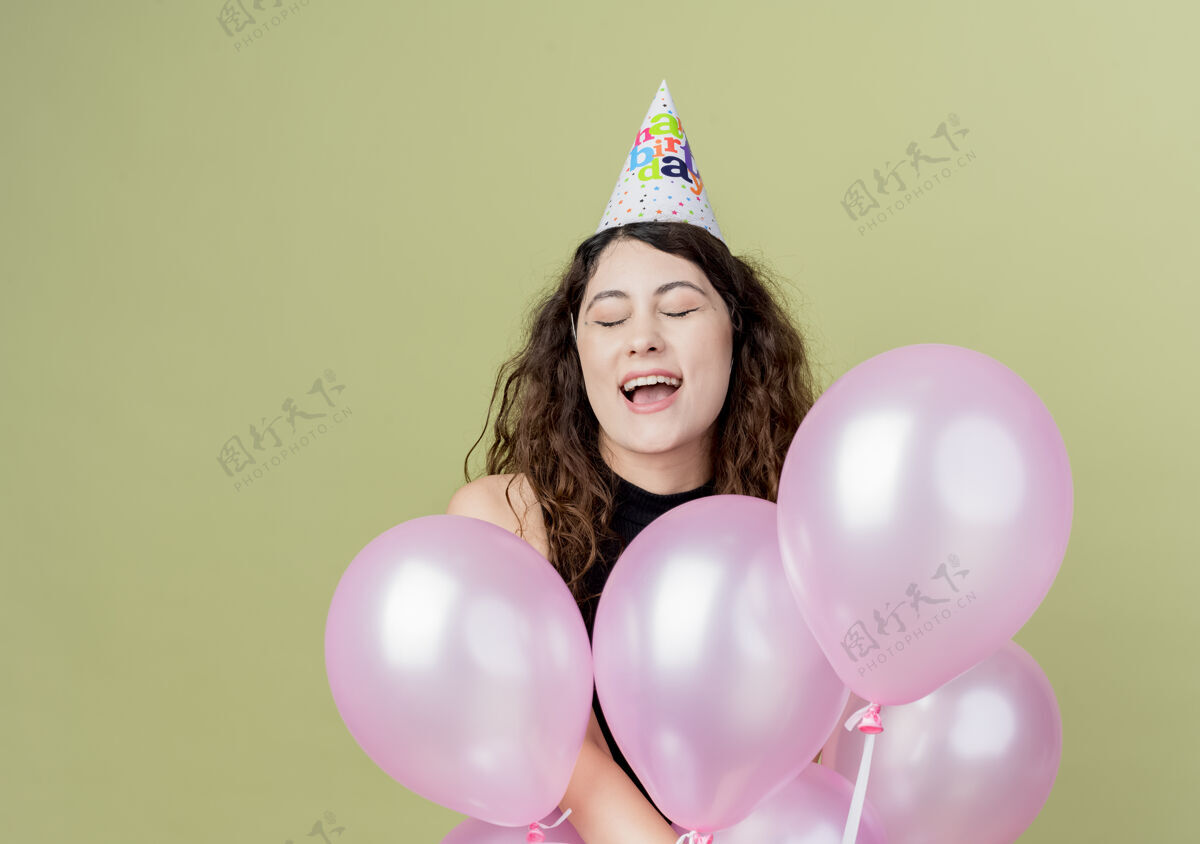 光年轻漂亮的女人 卷发 戴着节日帽 手里拿着气球 快乐而兴奋的生日派对概念站在光墙上抱着气球年轻