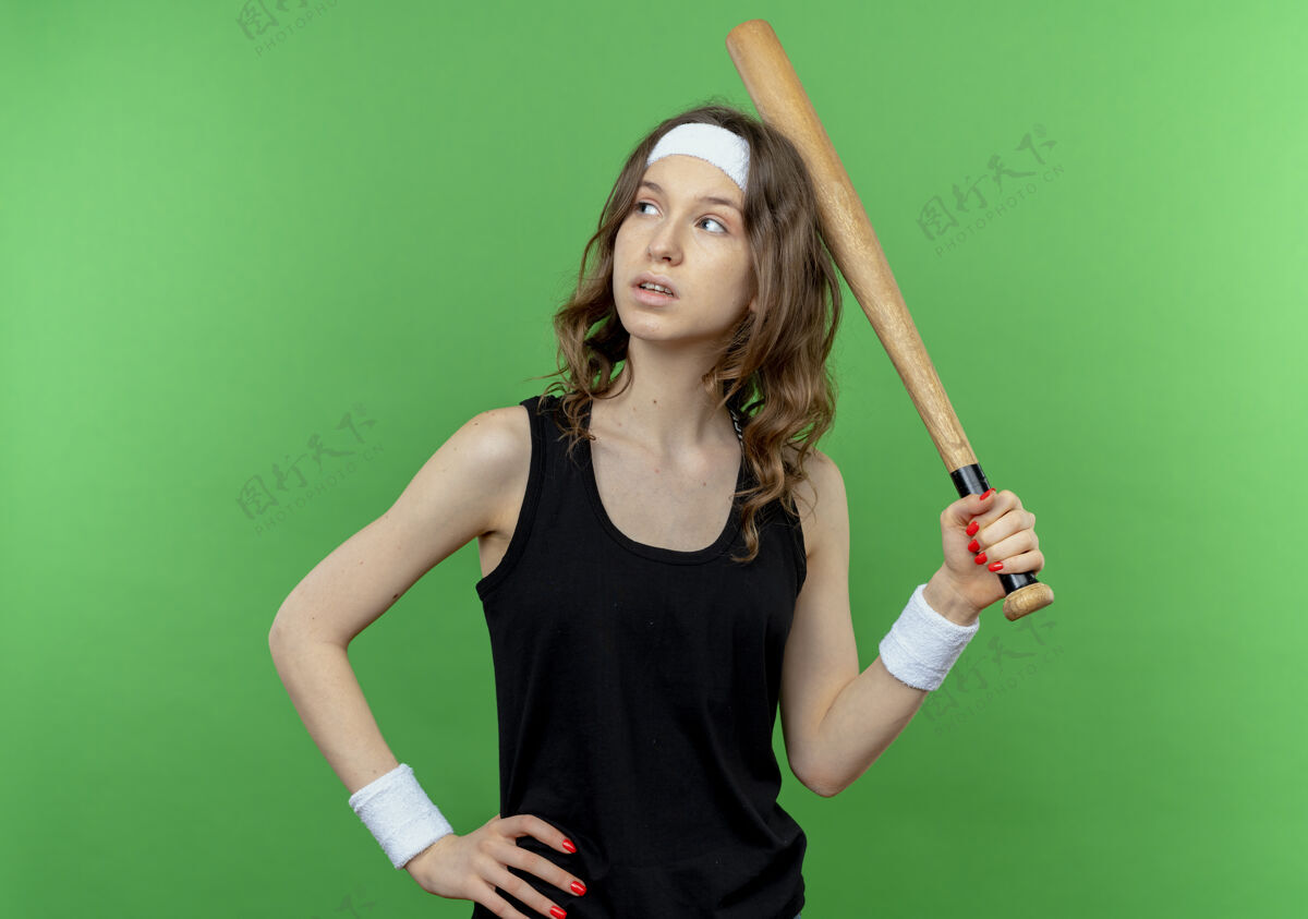 蝙蝠身穿黑色运动服的年轻健身女孩 头上戴着棒球棒 站在绿色的墙上 听着头在一旁沉思的表情年轻运动装表情