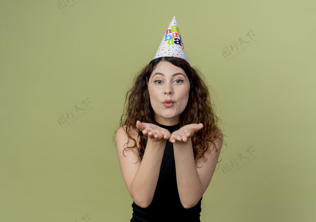 轻戴着节日帽的卷发美女站在轻墙上 在生日派对前用手吹着吻卷发站吹