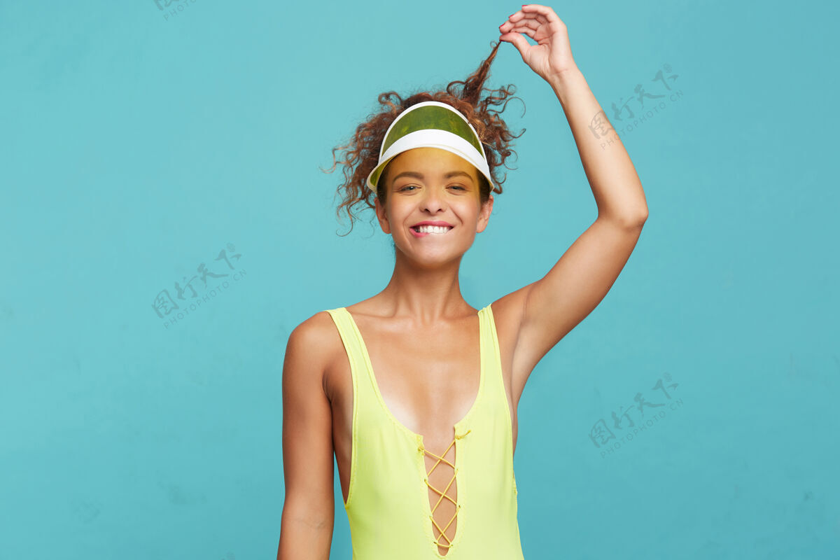 女士摄影棚拍摄的年轻可爱的红发女子拉着她的卷发 举起手 咬着内衣 同时积极地看着相机 站在蓝色的背景上高兴情绪帽子