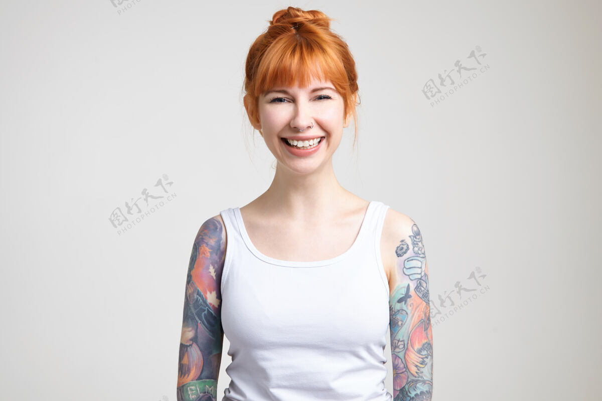 微笑摄影棚照片年轻漂亮的红发纹身女子与发髻的发型看着幸福的相机与广泛的微笑 而站在白色的背景白皙鼻子情绪