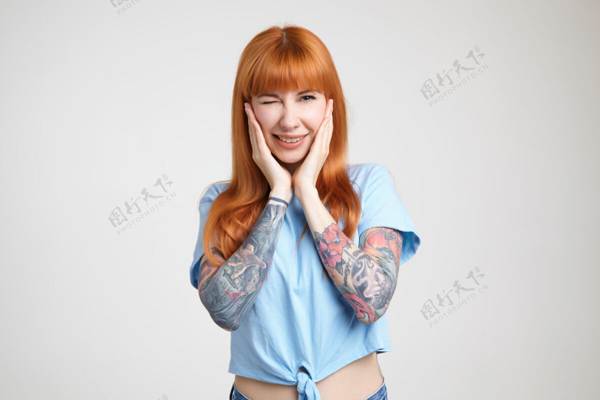 白皙快乐的年轻可爱的红发纹身女性举起双手捧着脸 高兴地对着镜头眨眼 站在白色的背景下墨水戒指眼睛