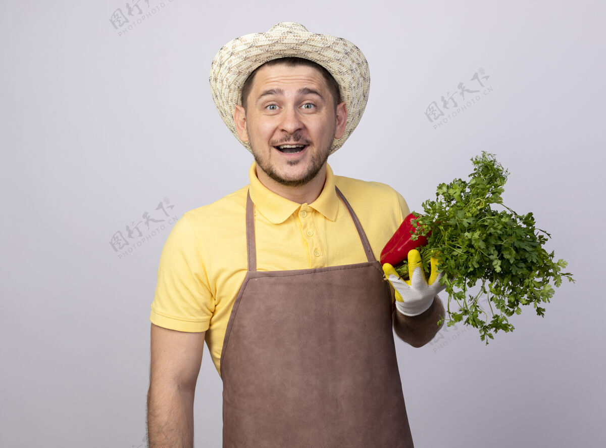 看年轻的园丁穿着连体衣 戴着帽子 戴着工作手套 手里拿着红甜椒和新鲜的草药 微笑着花园拿着帽子