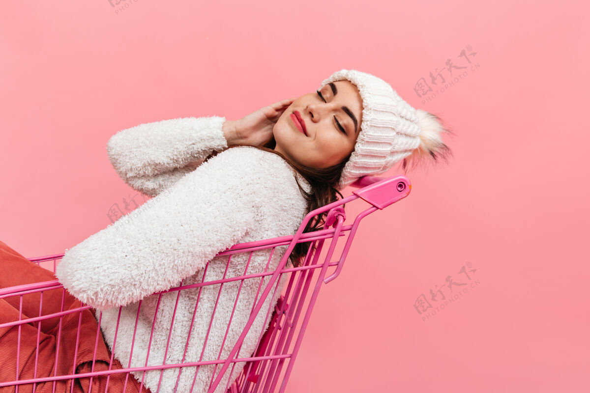 模特安静的闭着眼睛的女孩在粉红色的手推车里摆姿势戴着暖和的帽子和毛衣的女士的肖像享受微笑超市手推车