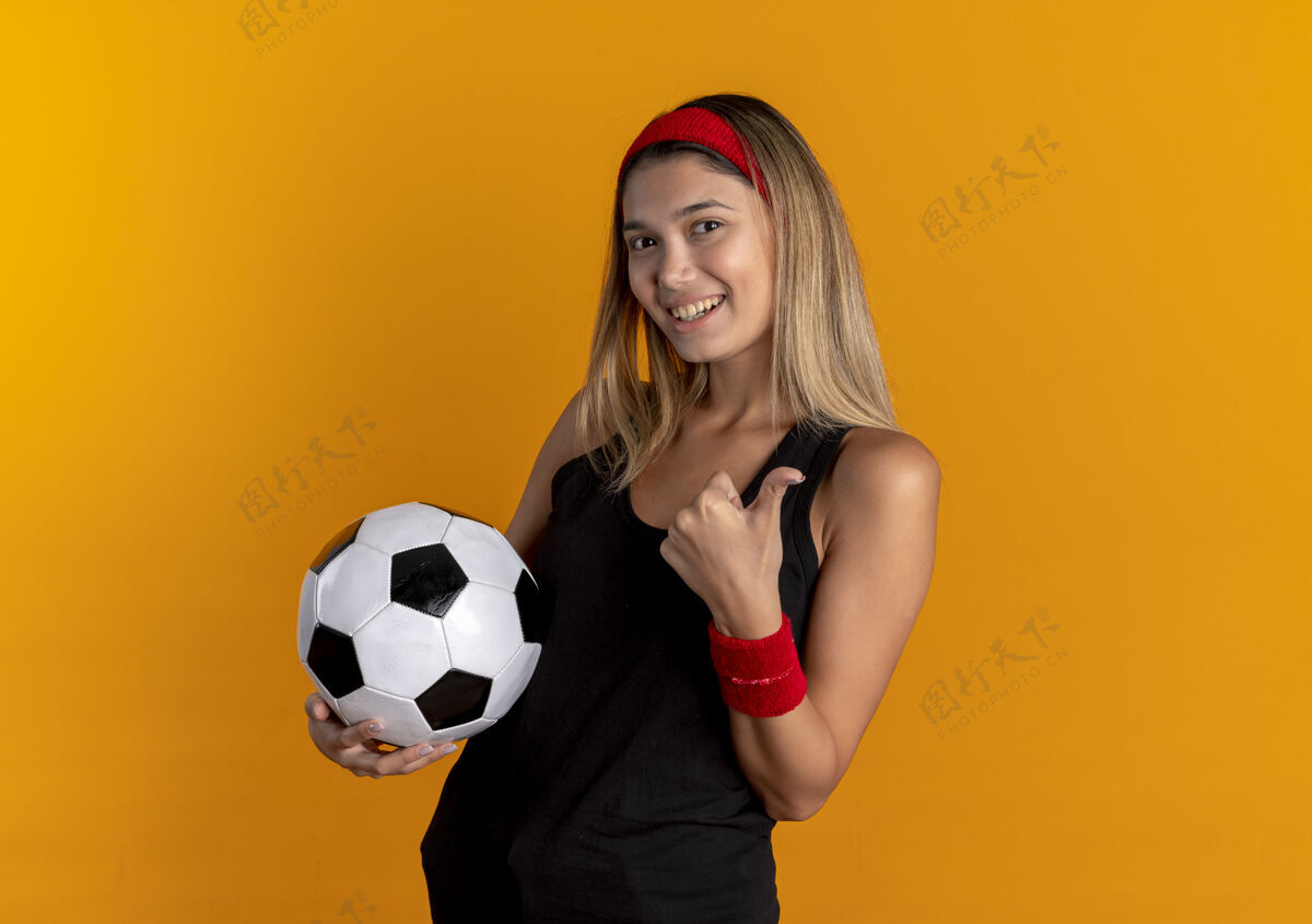运动装身穿黑色运动服 头戴红色头巾 手持足球 微笑着竖起大拇指的年轻健身女孩站在橙色的墙上健身年轻头带