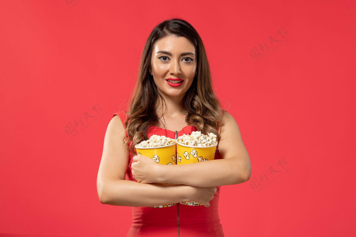 封面女郎正面图年轻女性手持爆米花包 微笑着站在红色的表面上举行包装电影