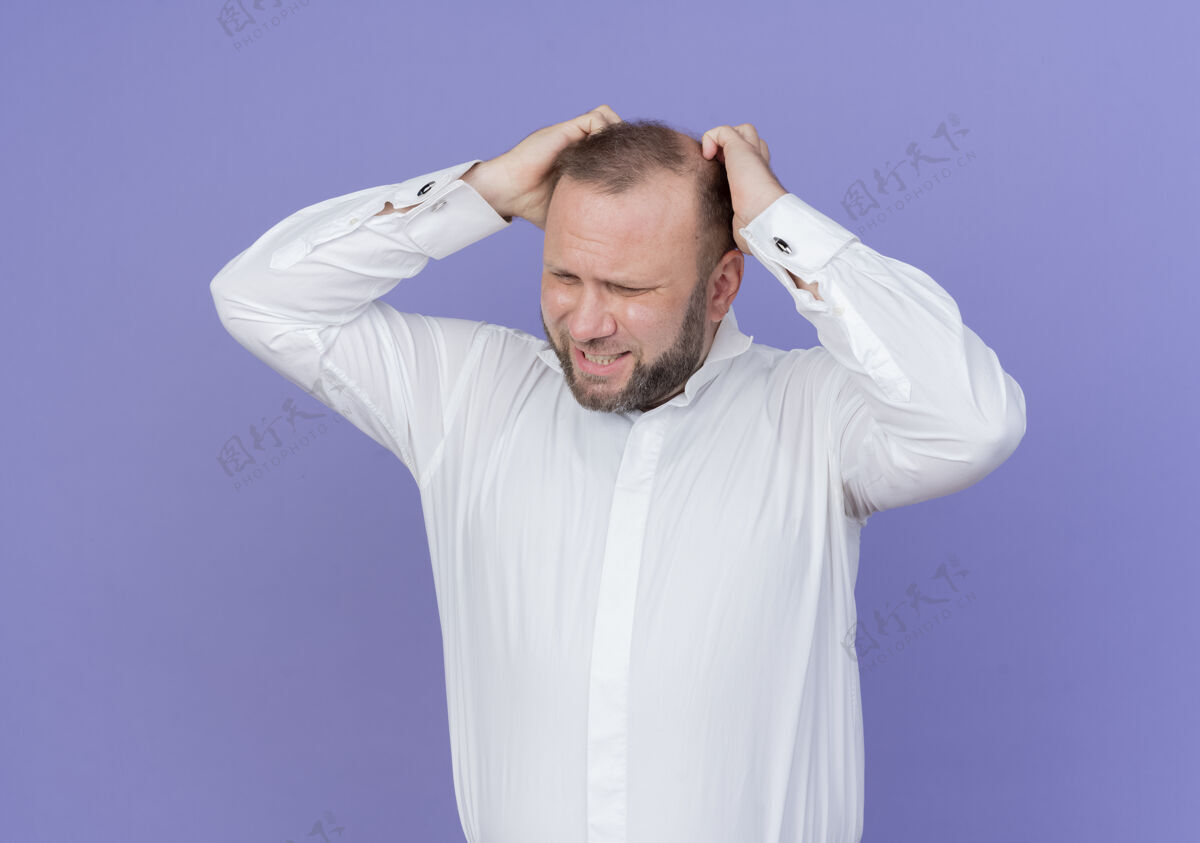 沮丧心烦意乱的胡子男人穿着白衬衫摸着他的头 站在蓝色的墙上感到沮丧摸沮丧男人
