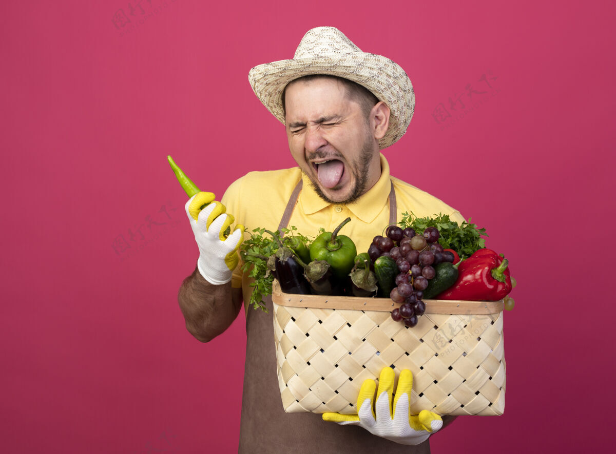 蔬菜年轻的园丁戴着工作手套 穿着连体衣 戴着帽子 手里拿着装满蔬菜的板条箱 嘴里吐着绿辣椒酱 表情很恶心舌头花园板条箱