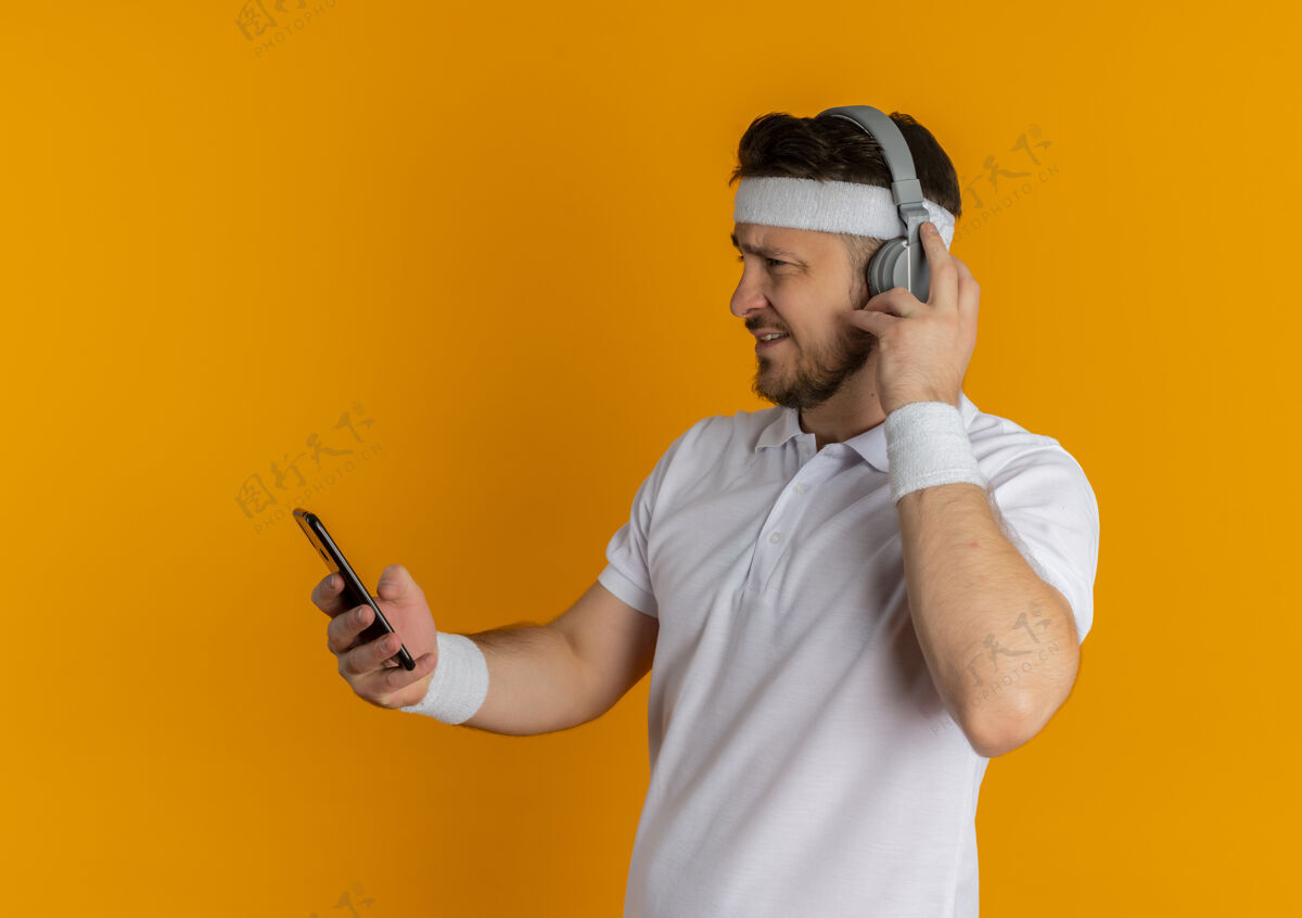 健身一个穿着白衬衫 戴着头带和耳机的年轻人站在橙色背景上看着手机屏幕上搜索音乐衬衫搜索白色
