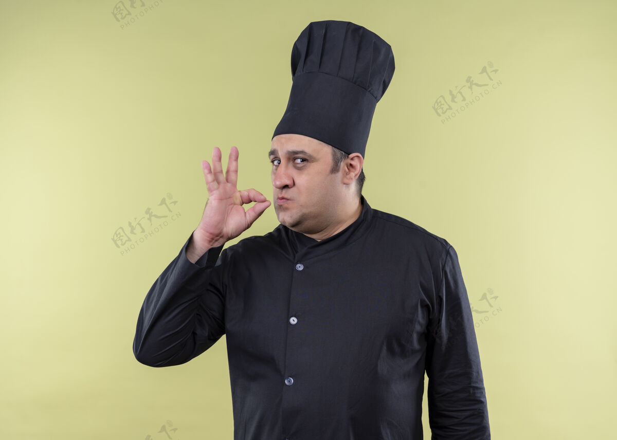 男性男厨师身穿黑色制服 头戴厨师帽 站在绿色背景上 展示美味的标志烹饪制服美味