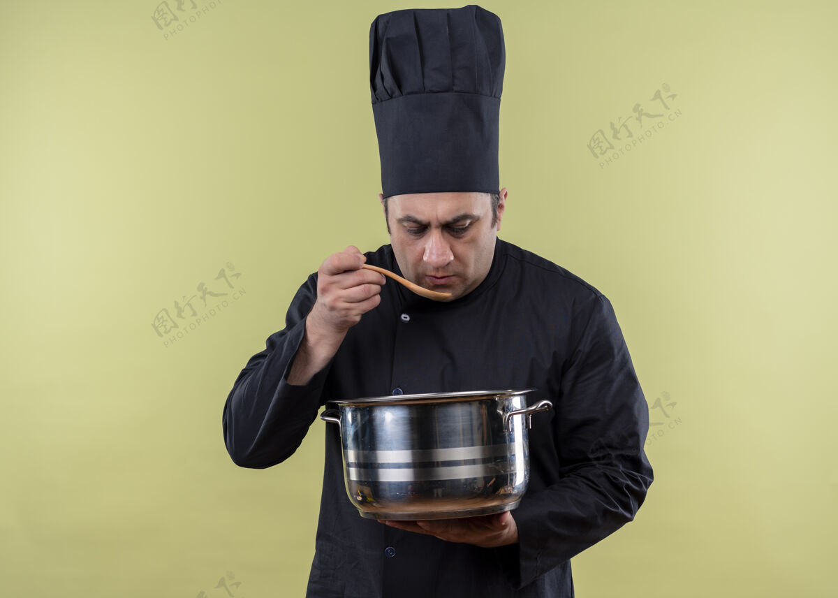 勺子男厨师身穿黑色制服 头戴厨师帽 手持平底锅 在绿色背景上用木勺品尝食物站着食物平底锅