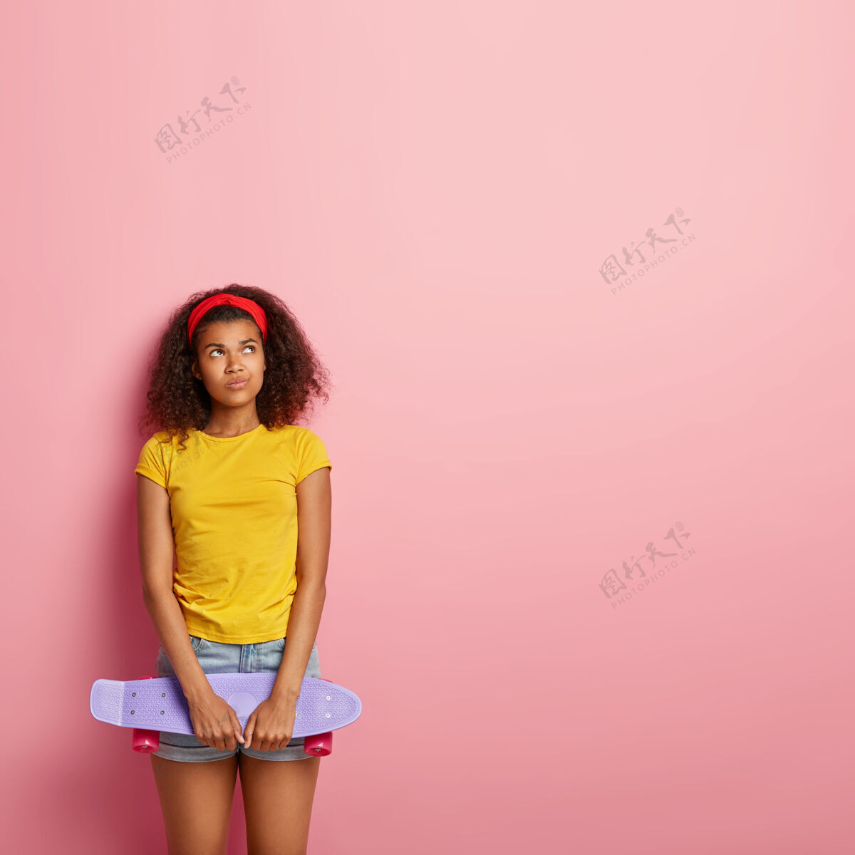 头带身材苗条漂亮的非裔美国女人的垂直镜头有着体贴的不满表情 拿着滑板滑冰 戴着红色的头带 黄色的t恤和牛仔短裤 模特们在玫瑰色的墙上人魅力清爽