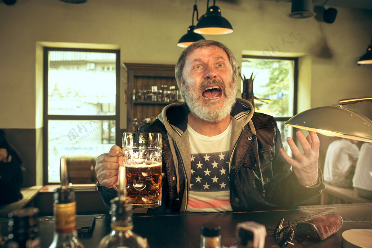 球迷大胡子男人在酒吧喝酒 看电视上的体育节目享受我最喜欢的酒和啤酒坐在桌上喝啤酒的男人足球或体育迷人类情感的概念乐趣啤酒老年人