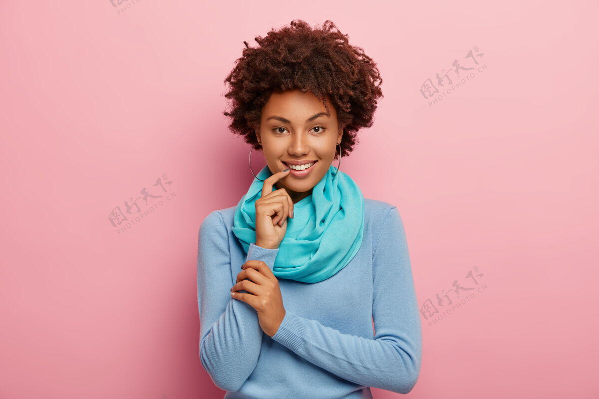 表情半身镜头 可爱的美国黑人女子 清爽的头发 穿着蓝色套头衫 丝巾 手指靠近嘴唇 看起来神秘地孤立在粉红色的墙上情感概念肖像粉色模特