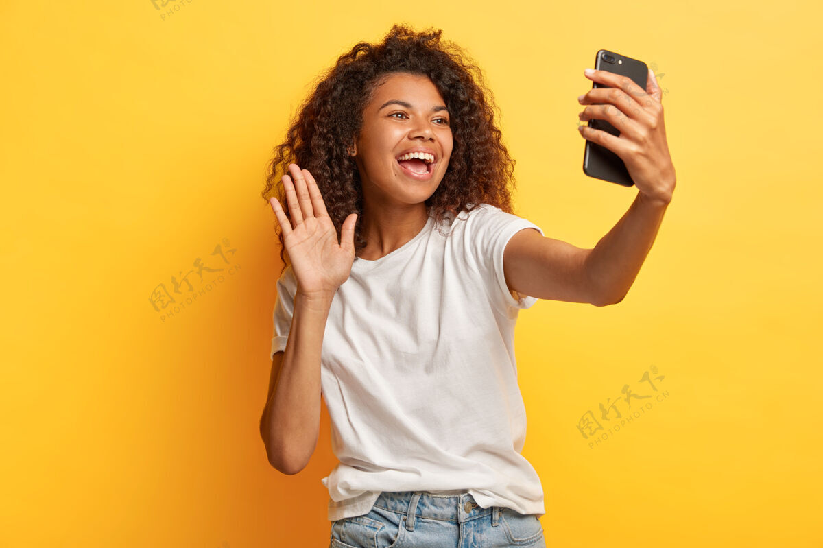 黑色快乐的黑皮肤快乐的女人 非洲发型 手持现代手机在面前 在镜头中挥舞手掌 打视频电话 穿着休闲装室内爽快牙关