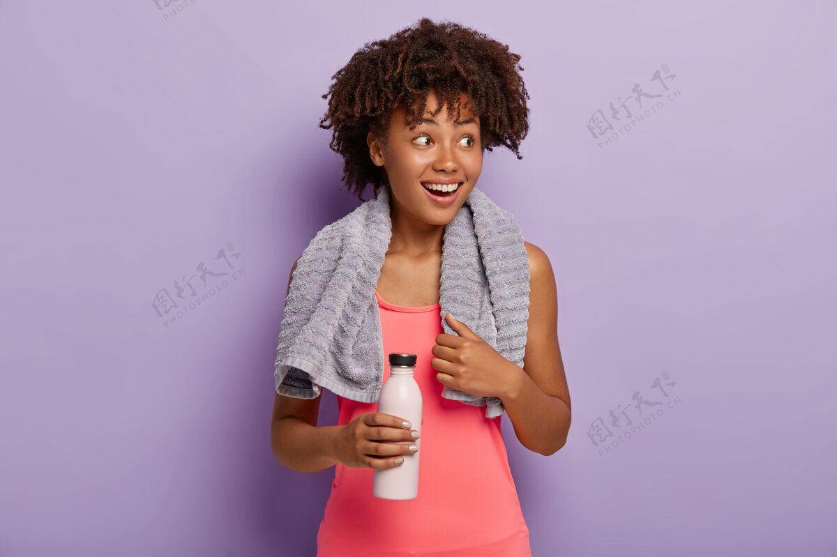 积极照片中快乐的黑皮肤女运动员留着非洲式发型 微笑着看向一边 穿着粉色上衣 端着瓶子 在训练中口渴时喝水 在室内锻炼欢呼休息锻炼