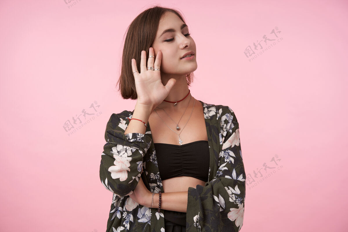 发型可爱的年轻积极的棕色头发的女人穿着花衬衫和黑色上衣 展示了抬高的手掌和微笑 而在粉红色的墙上闭着眼睛摆姿势自然提高手