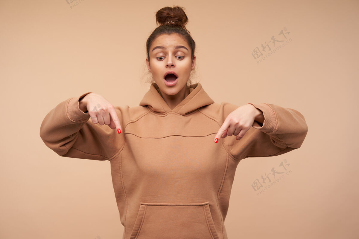 衣服激动的年轻漂亮的棕色头发的女人 穿着运动服 举起双手 用食指指着下面 站在米色的墙上站女性摩卡
