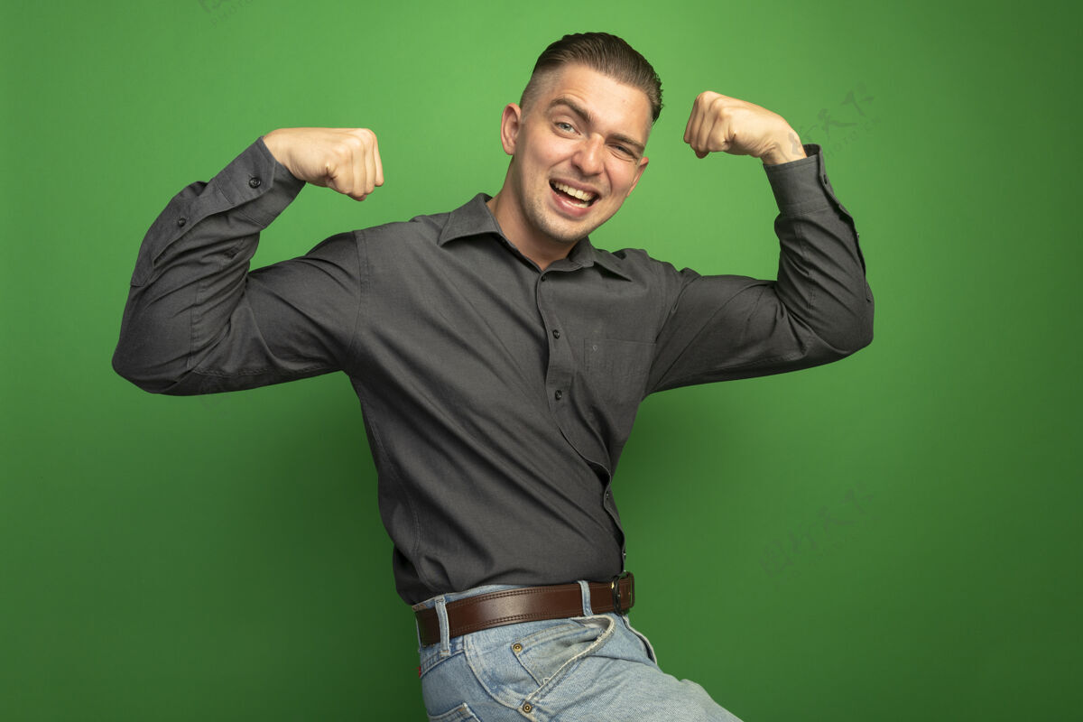 人身着灰色衬衫的年轻帅哥举起拳头 高兴而积极地展示了站在绿色墙壁上的肱二头肌和力量赢家概念灰色绿色力量